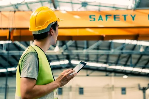 A warehouse worker in a hard hat and hi-vis vest checking a tablet beneath a safety sign